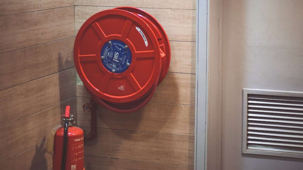 A red fire hose reel is mounted on a wooden wall next to a closed door, echoing the punctual precision of a chimney repair in Fairfield County, CT. Below the reel, a red fire extinguisher rests on the floor. The setting appears to be an indoor area, possibly a hallway or corridor.