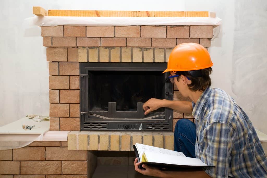 A construction worker in an orange hard hat and plaid shirt is inspecting a newly installed brick fireplace, likely assessing the chimney cap details. He points at the glass door while consulting a manual, ensuring everything meets Chimney Repair standards in Fairfield County CT.
