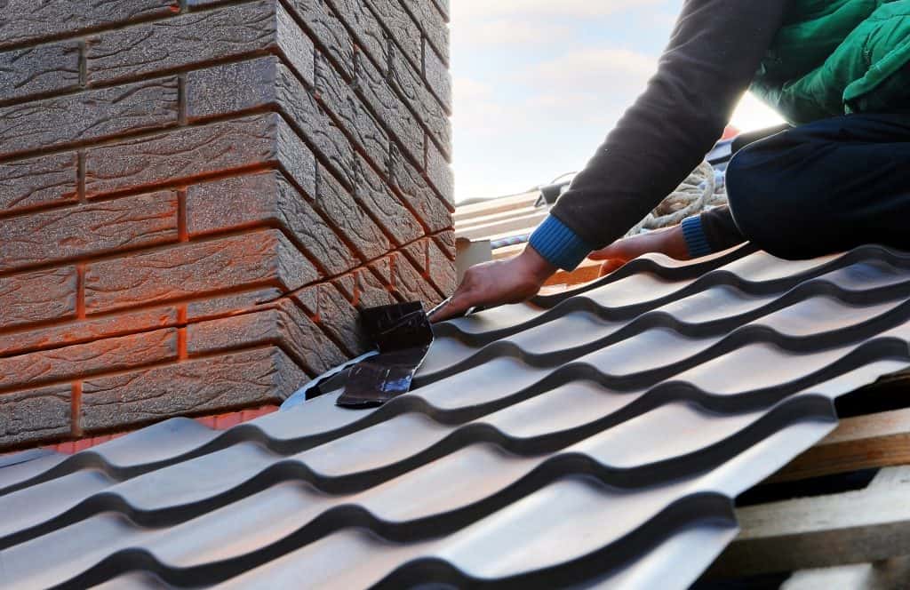 A person is expertly installing a metal roof around a brick chimney, ensuring a snug fit with precise flashing. The wavy-patterned roof gleams under the partly cloudy sky, showcasing skilled craftsmanship. Whether it's roofing or chimney repair in Fairfield County CT, attention to detail is key.